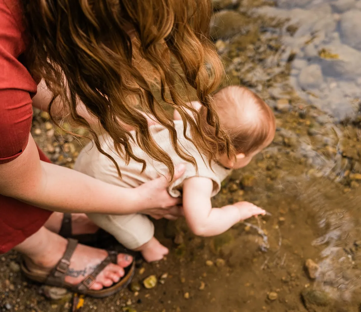 A parent steadies their baby as the baby reaches for the water at a shallow creek.