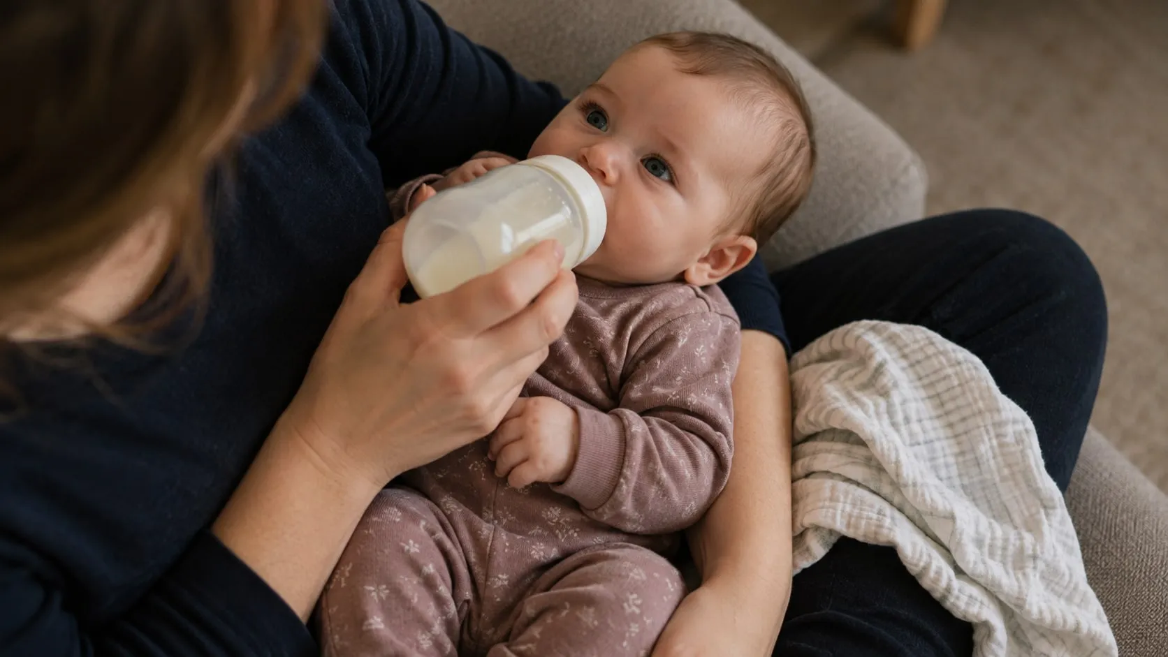 A parent feeds their baby a bottle on the couch, a muslin cloth beside them.