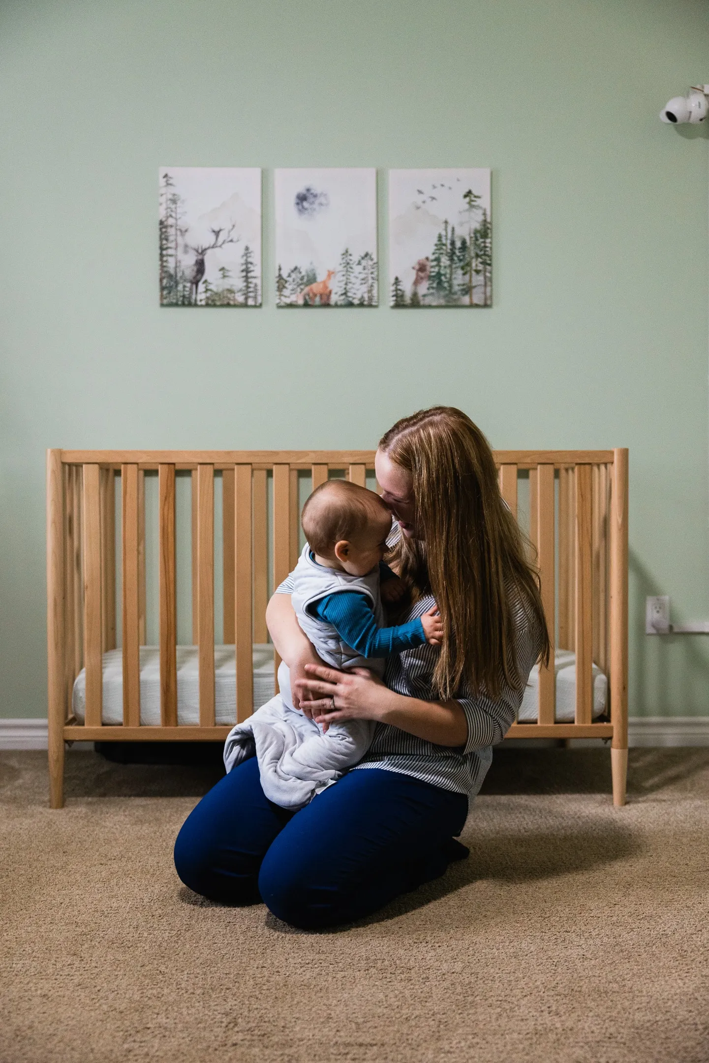 Nicole Graff-Rowe kneels beside a crib in a nursery, hugging her baby.