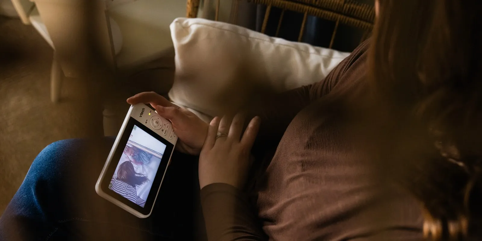A mother sits curled up in a wicker chair, looking at a photo on her phone.