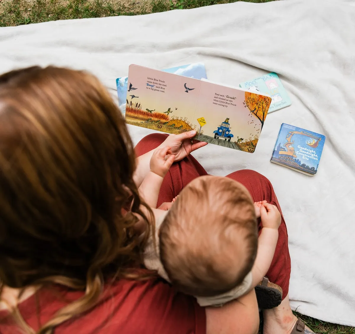 A mother sits on a blanket on the grass, reading a picture book to her baby in her lap.